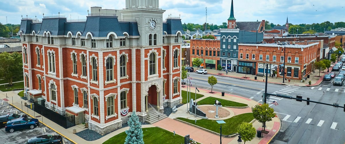 A historic building with a clock tower, surrounded by a city street with cars and a church with a spire.