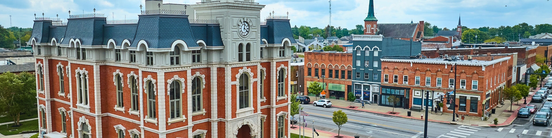 A historic building with a clock tower, surrounded by a city street with cars and a church with a spire.