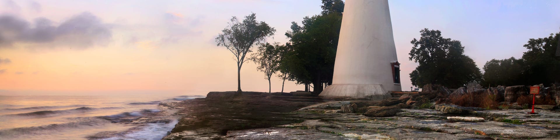 A lighthouse with a green light atop, situated on rocky terrain near the ocean with trees in the background.