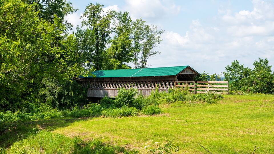 A wooden barn with a green roof surrounded by lush greenery and trees.