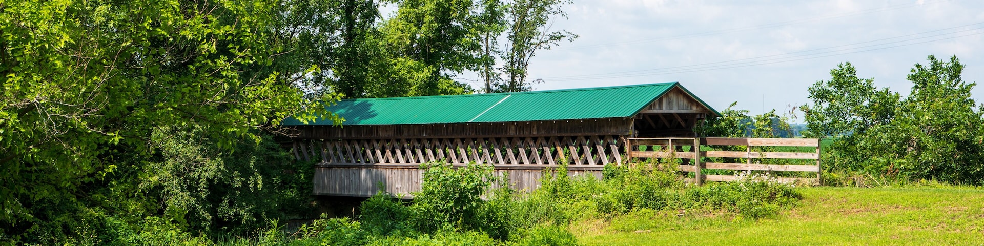 A wooden barn with a green roof surrounded by lush greenery and trees.