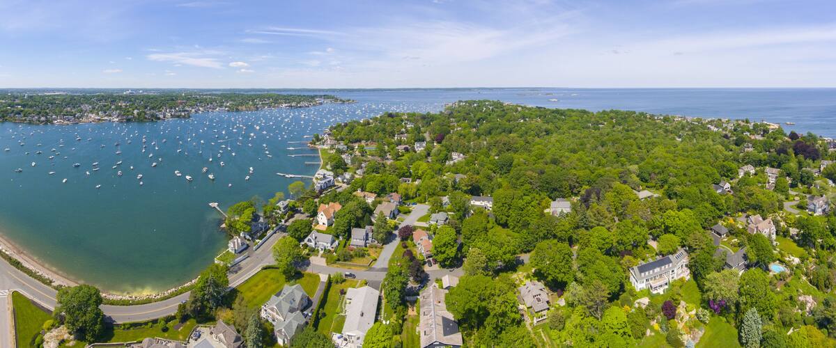 An aerial view of a coastal town with a marina filled with boats, surrounded by lush greenery and residential areas.