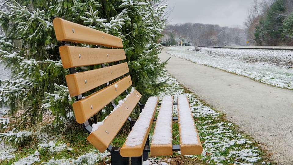 A wooden bench with snow on its backrest and seat, positioned beside a snow-covered path with trees in the background.