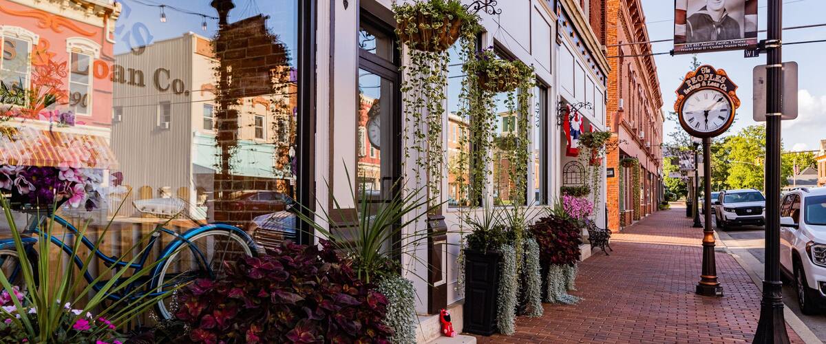 West Union, United States - 13 August 2025: View of a quaint street with brick buildings, flower-filled planters, and a vintage clock casting shadows on the red brick pavement.