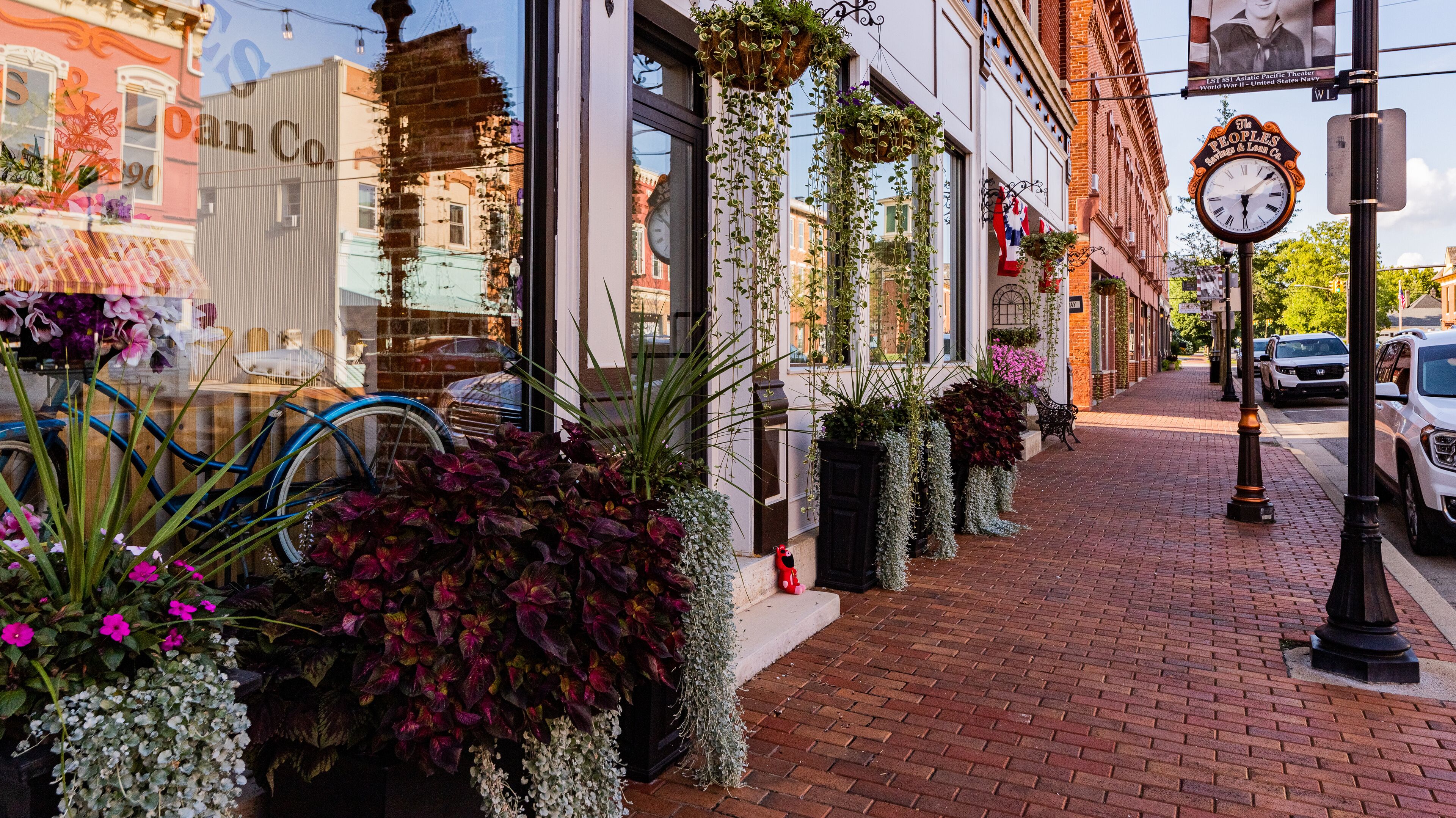A brick-paved street with hanging plants, a bicycle, and a vintage clock.