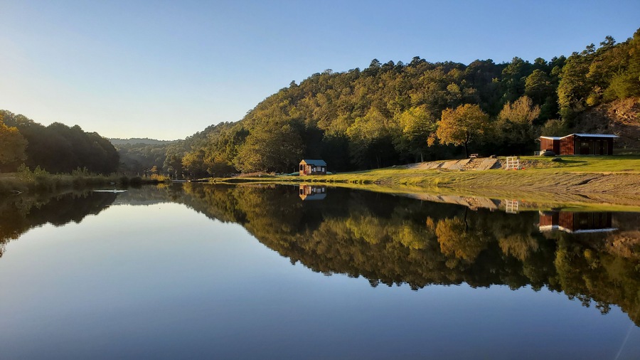 Un lac paisible, avec une maison en bois et une remise situés sur la rive droite, le tout entouré d’une forêt dense.