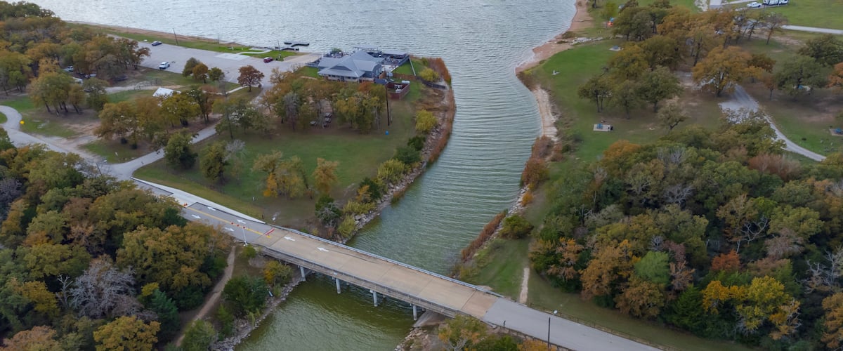 A bridge over a river, surrounded by trees and a parking area.