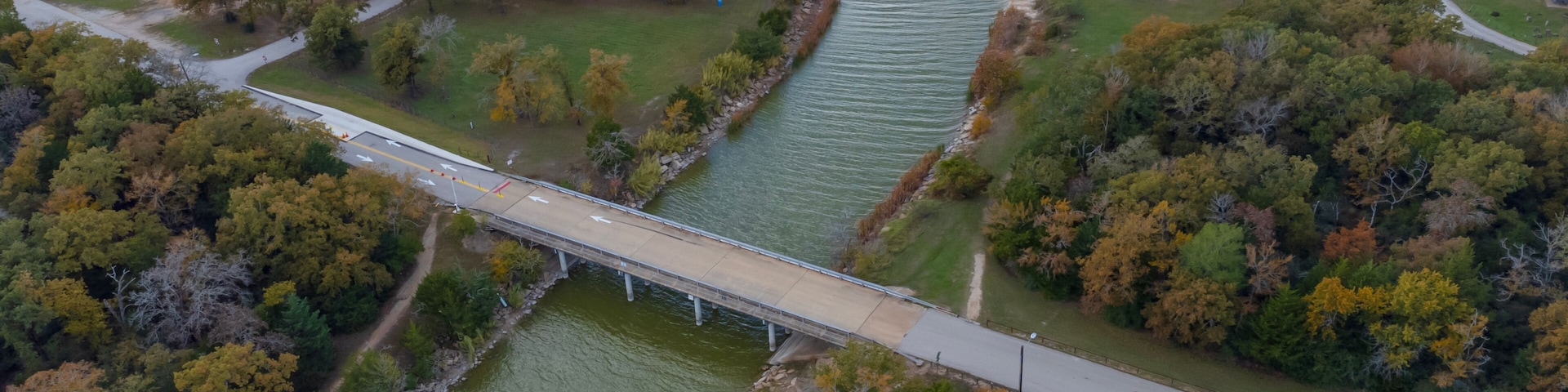 A bridge over a river, surrounded by trees and a parking area.