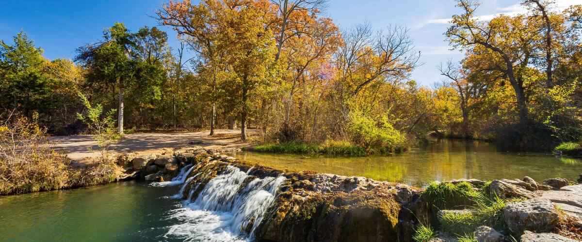A river with a waterfall, surrounded by autumn-colored trees and a clear blue sky.