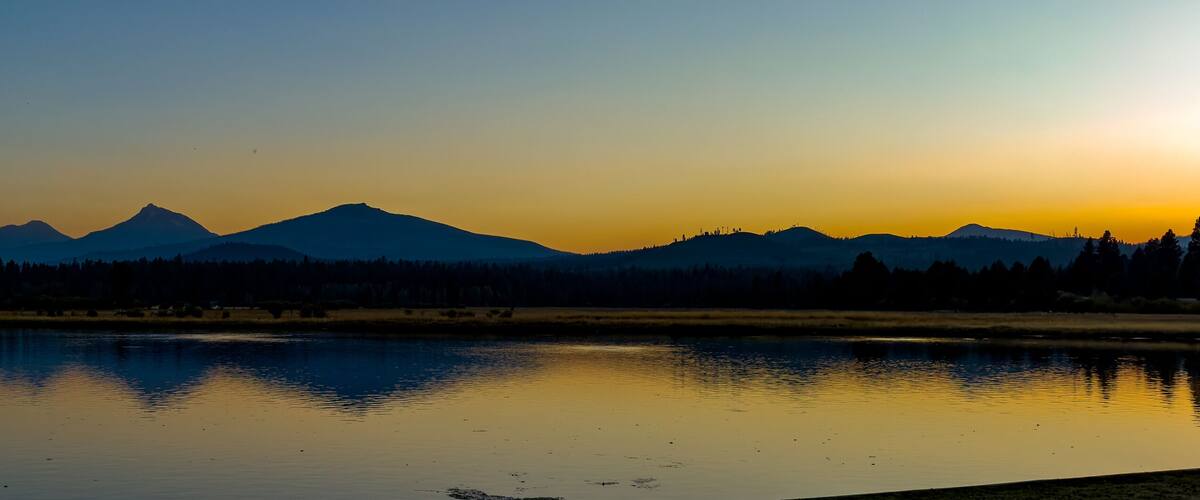 A panorama image of the Three Sisters mountains and Broken Top reflected at sunset in Lake Thalarope on Black Butte Ranch near Sisters Oregon
