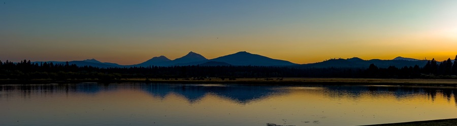 A panorama image of the Three Sisters mountains and Broken Top reflected at sunset in Lake Thalarope on Black Butte Ranch near Sisters Oregon