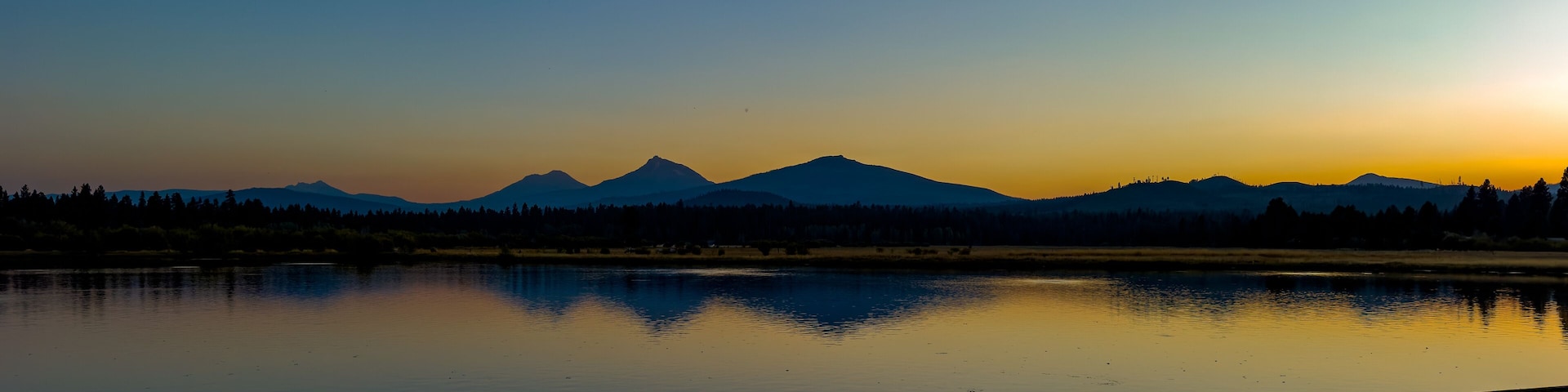 A panorama image of the Three Sisters mountains and Broken Top reflected at sunset in Lake Thalarope on Black Butte Ranch near Sisters Oregon