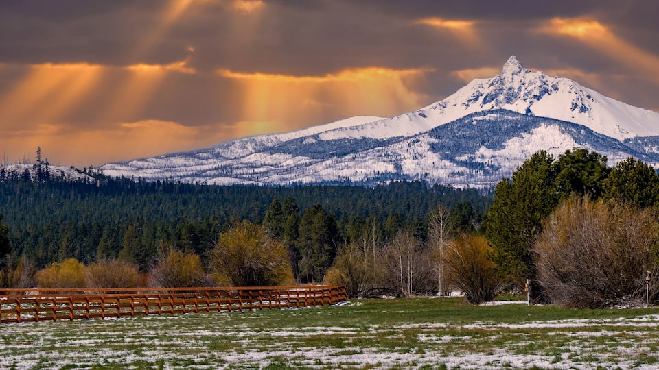 A snow-covered mountain with rays of sunlight breaking through the clouds, a wooden fence, and a forested valley.