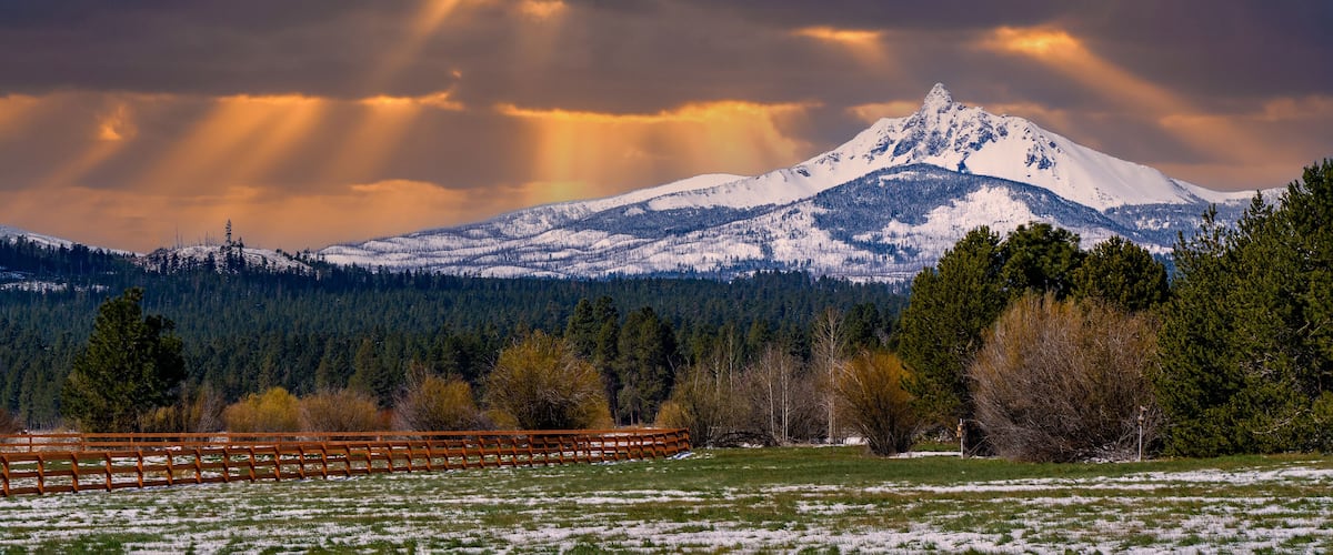 A snow-covered mountain with rays of sunlight breaking through the clouds, a wooden fence, and a forested valley.