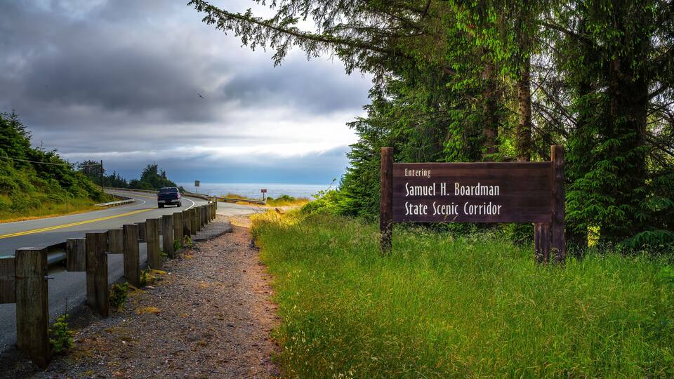 Road leading into Samuel H. Boardman State Scenic Corridor in Oregon with welcome sign and greenery.