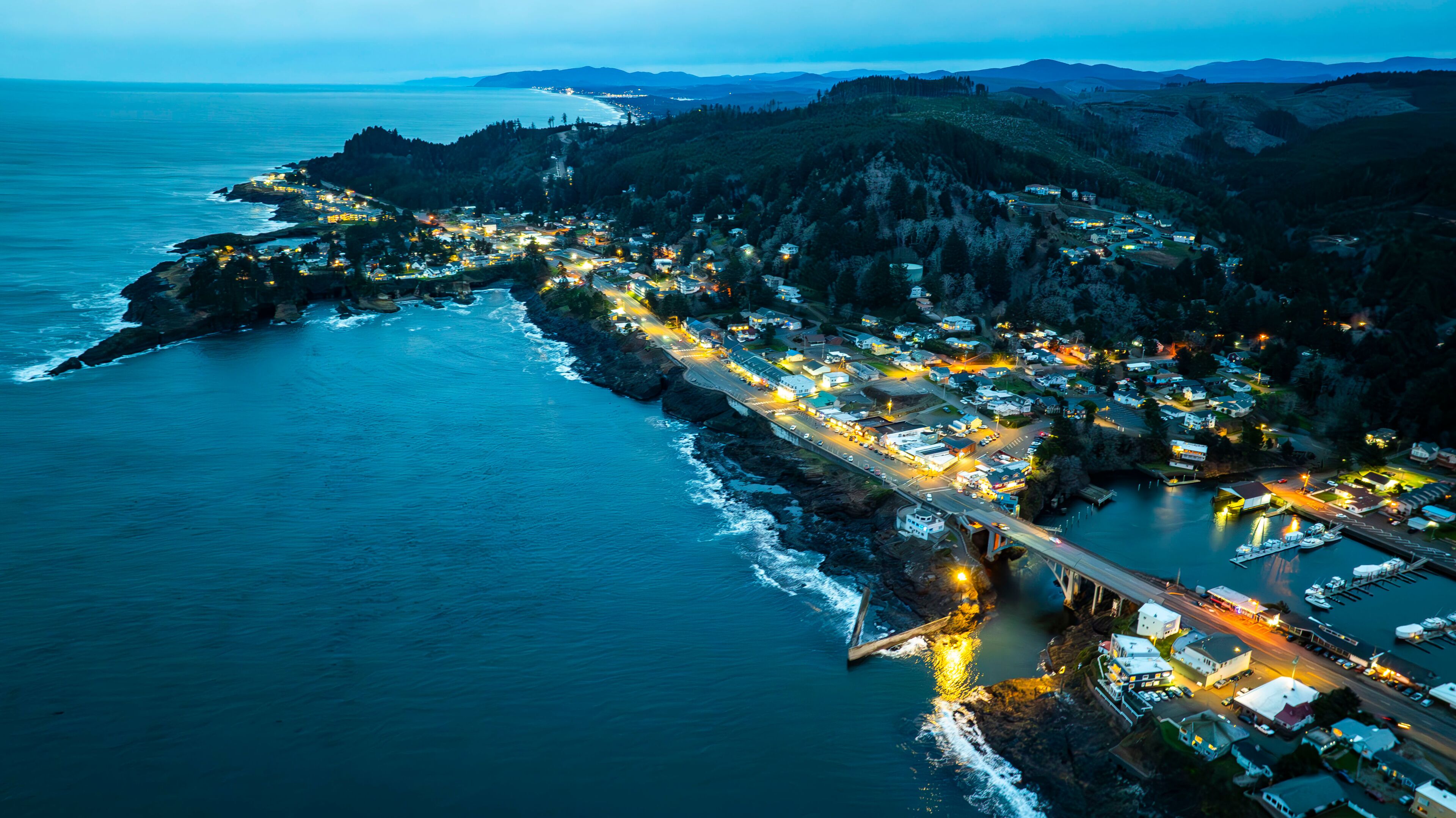 A coastal town at dusk with a bridge connecting the mainland to an island, surrounded by mountains and the ocean.