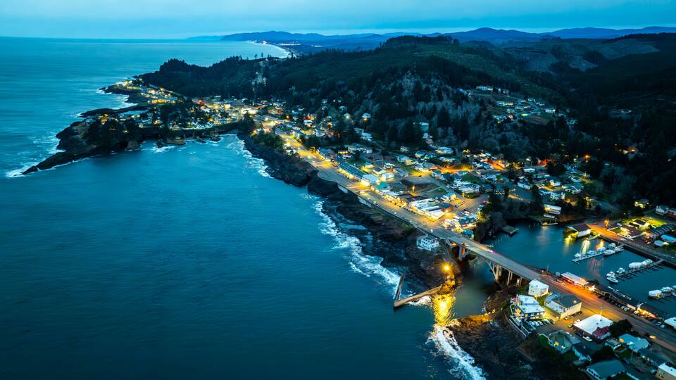 A coastal town at dusk with a bridge connecting the mainland to an island, surrounded by mountains and the ocean.