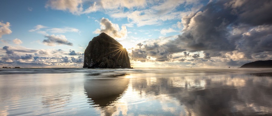 A calm beach with a large rock formation and the sun setting in the background.