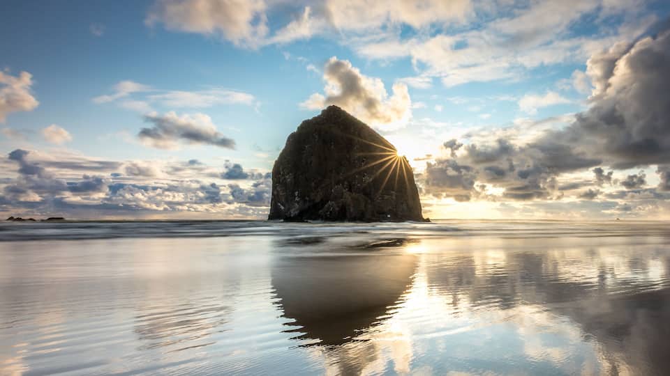 A calm beach with a large rock formation and the sun setting in the background.