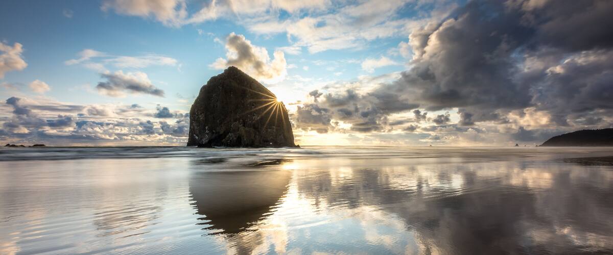 A calm beach with a large rock formation and the sun setting in the background.