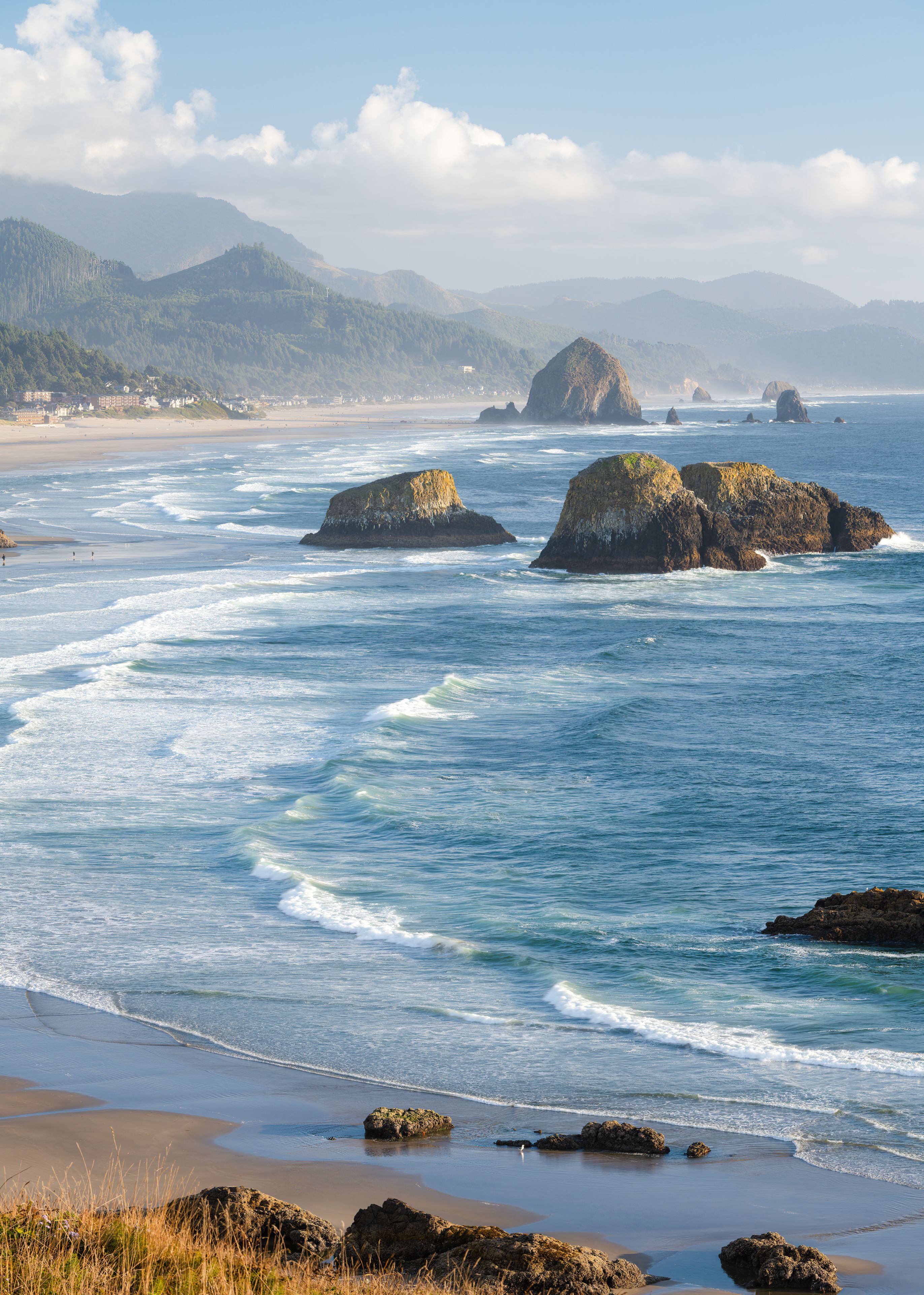 A coastal landscape with waves crashing onto a sandy beach, rocky outcrops, and distant mountains under a clear sky.