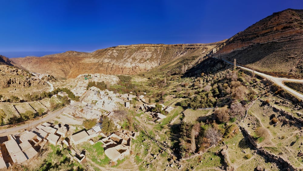 A panoramic view of a valley with a winding road, terraced fields, and a small settlement with buildings.