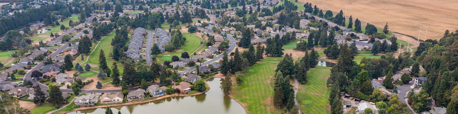 Aerial drone view of Staats Lake in Keizer, Oregon, near Salem, featuring a residential neighborhood, a golf course, and lush green landscapes under a clear blue sky