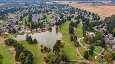 Aerial drone view of Staats Lake in Keizer, Oregon, near Salem, featuring a residential neighborhood, a golf course, and lush green landscapes under a clear blue sky