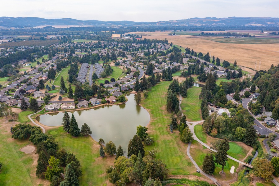 Aerial drone view of Staats Lake in Keizer, Oregon, near Salem, featuring a residential neighborhood, a golf course, and lush green landscapes under a clear blue sky