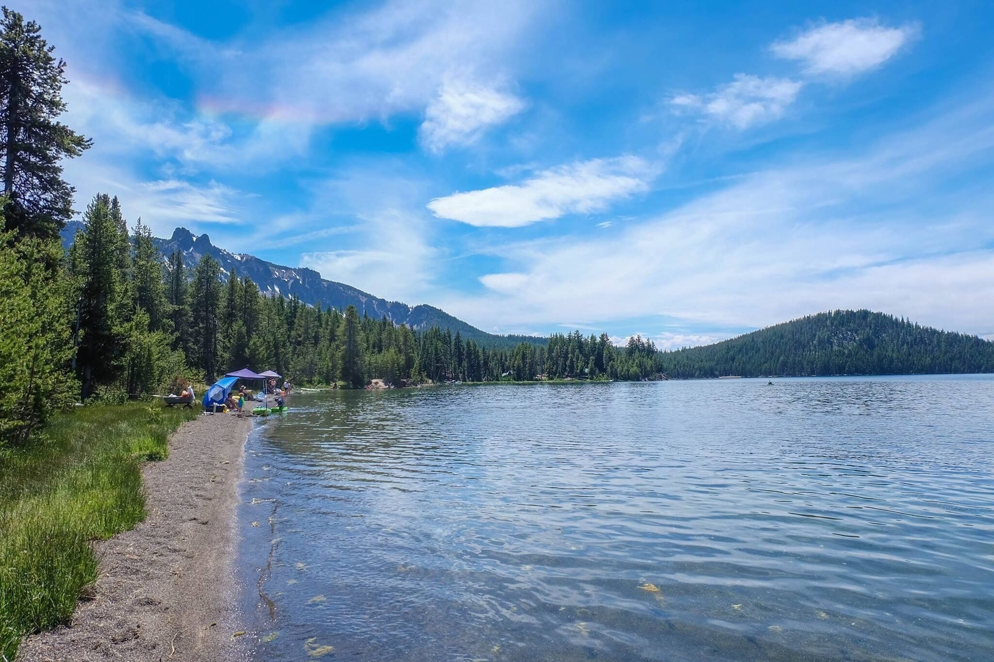 A calm lake with clear water, surrounded by dense forests and mountains under a blue sky with clouds.