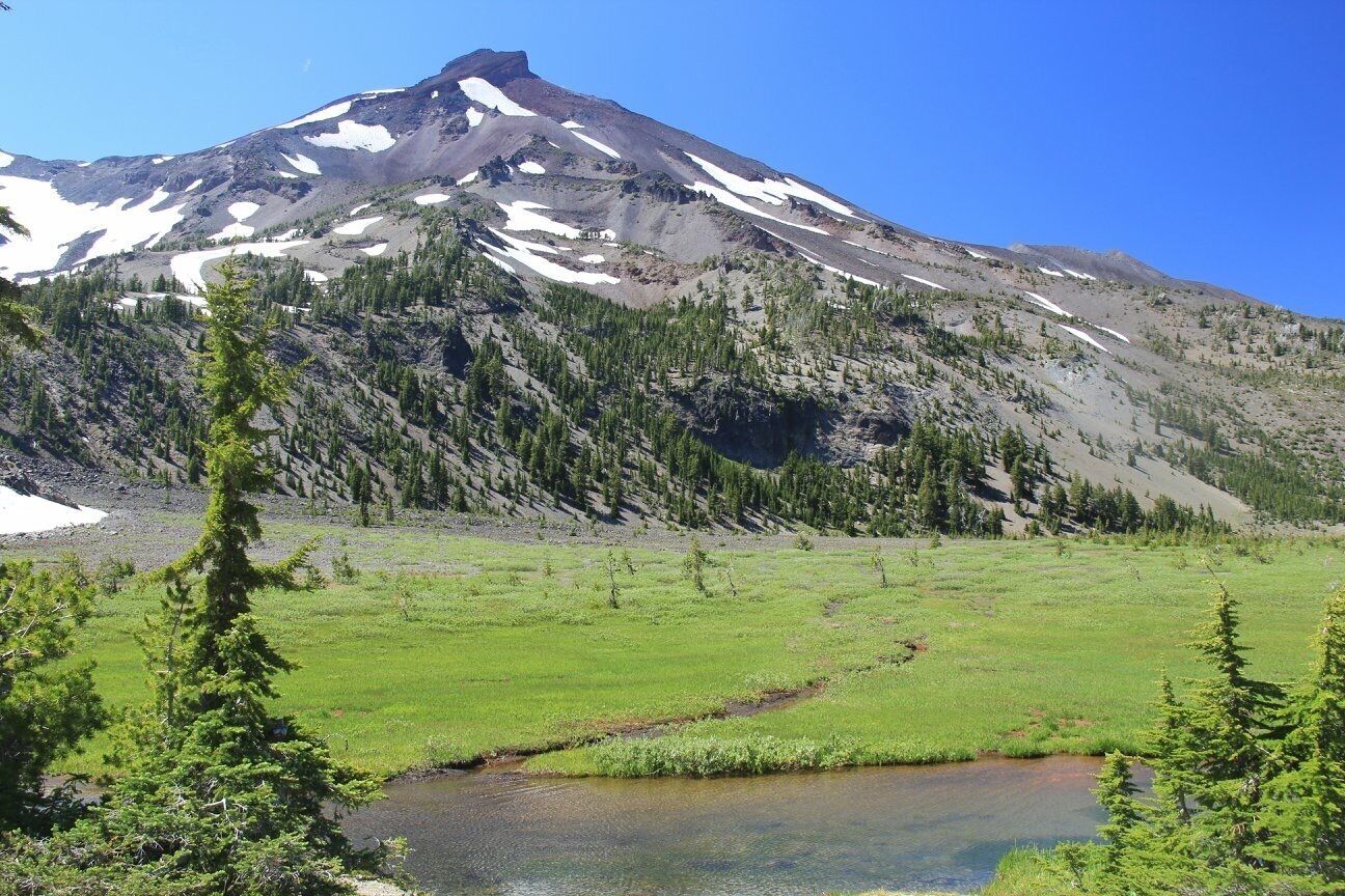 A mountain with snow patches, a green valley, and a river.