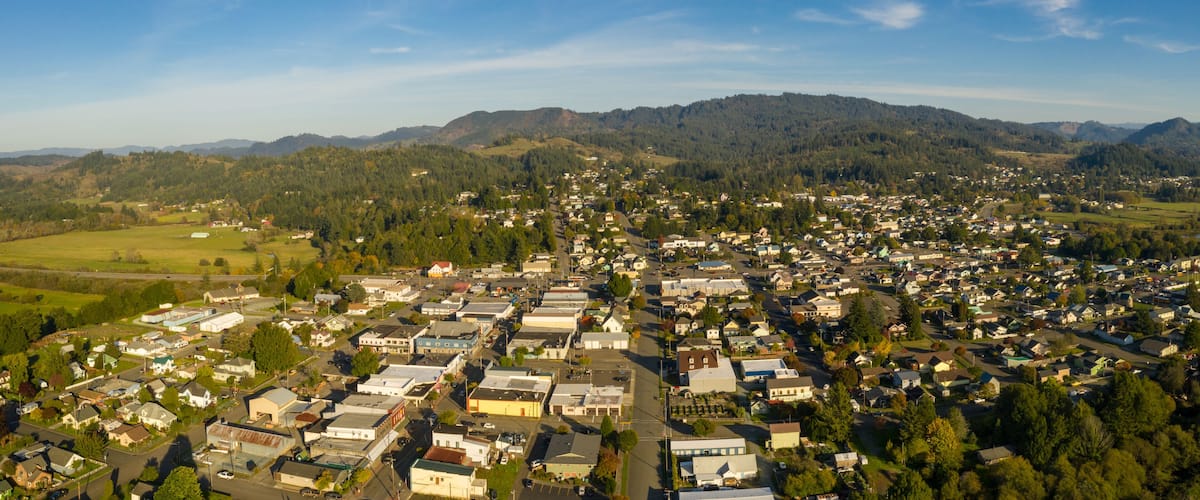 An aerial view of a town with a dense cluster of buildings, surrounded by green hills and mountains.