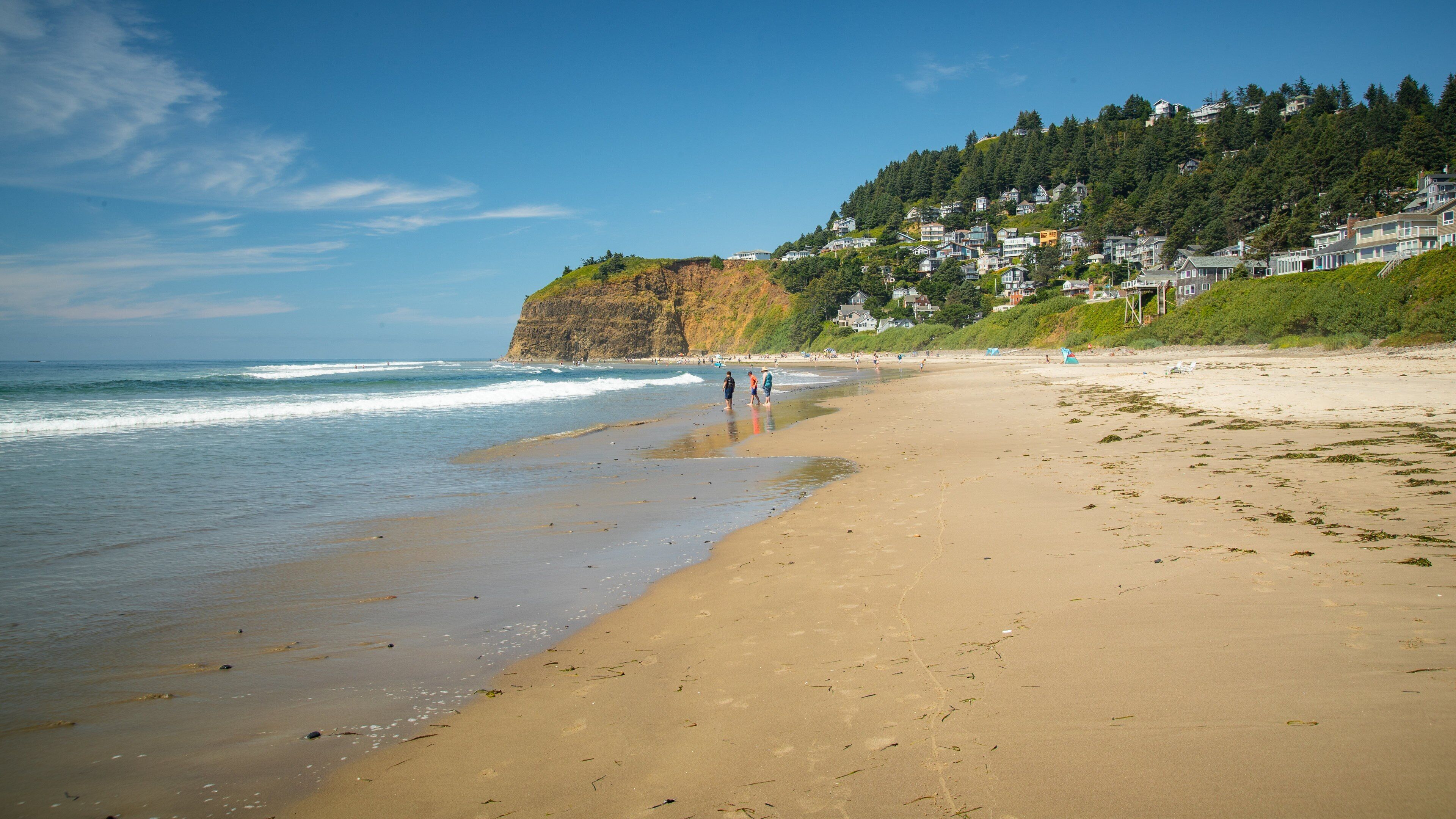 A sandy beach with waves, a cliff with houses, and a clear blue sky.