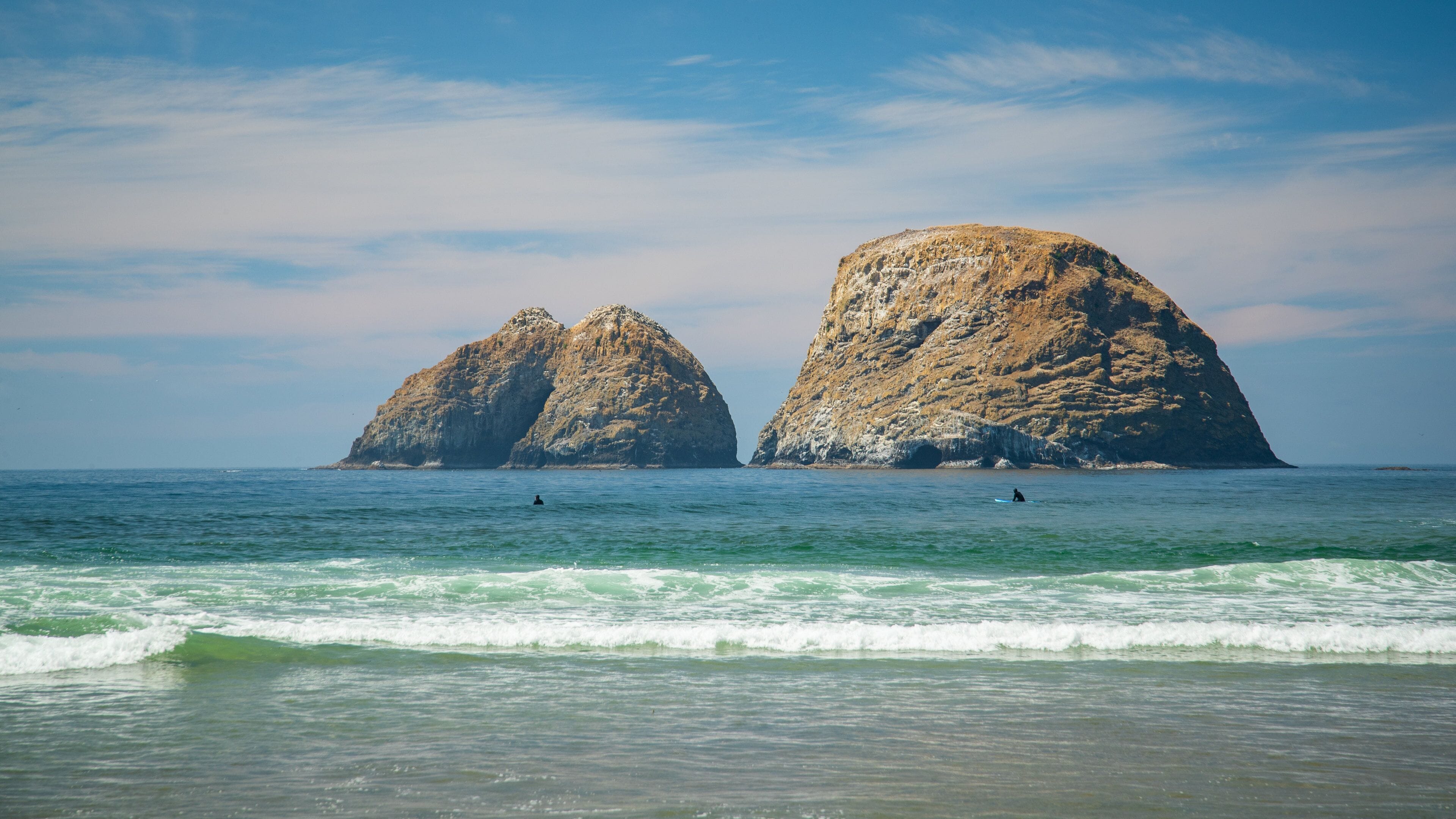Two large rock formations in the ocean with surfers in the water.