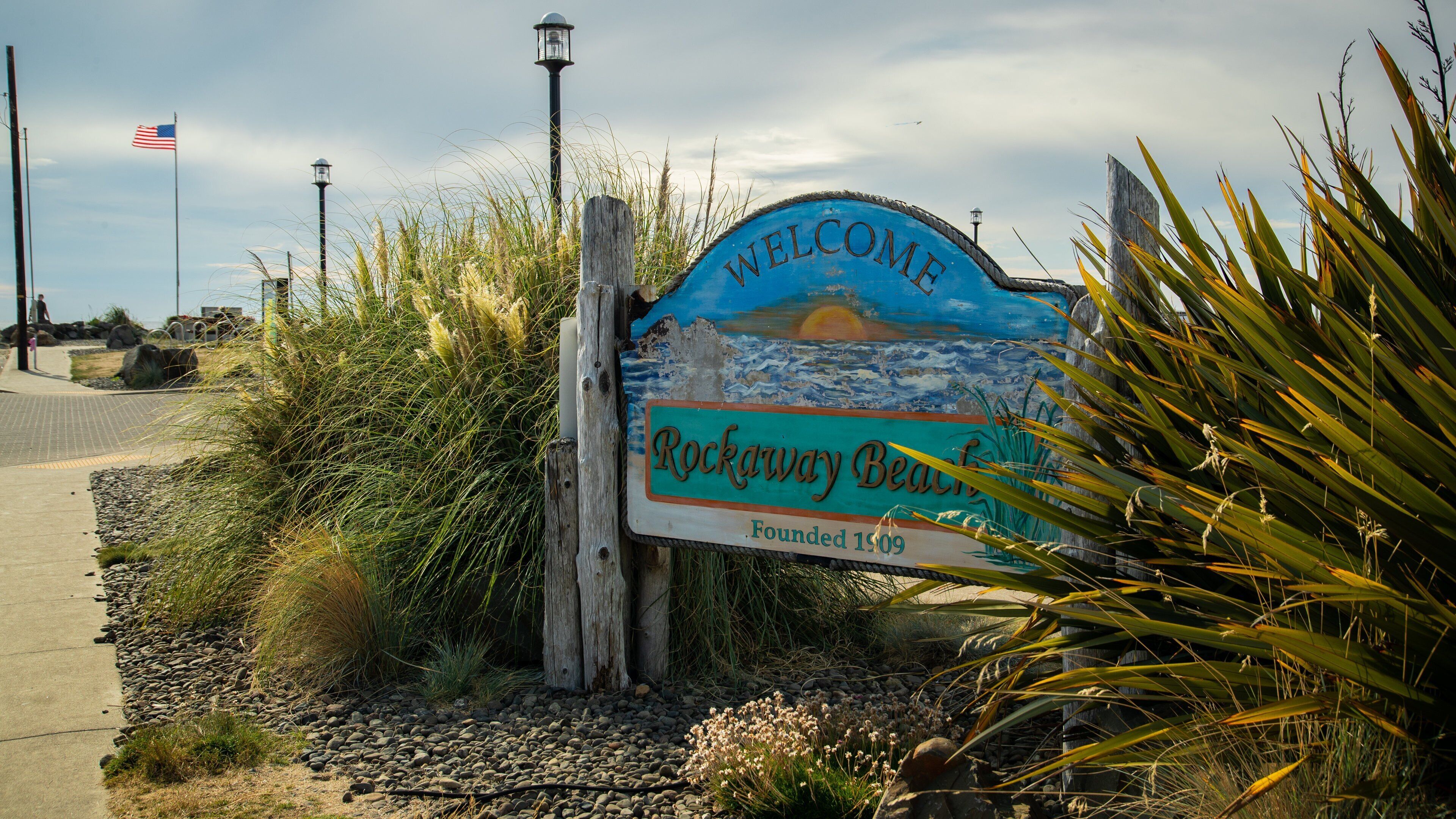 A welcome sign for Rockaway Beach with a coastal scene, an American flag, and a lamp post.