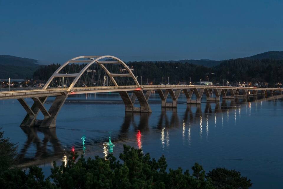 A modern bridge with a unique arch design spanning a calm river, surrounded by forested hills.