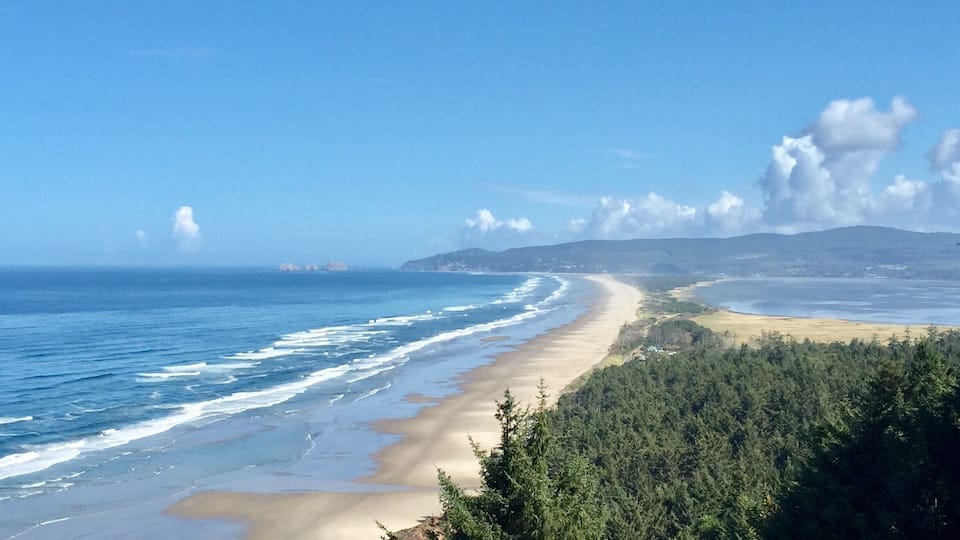 A coastal landscape with a sandy beach, ocean waves, dense forest, and distant hills.