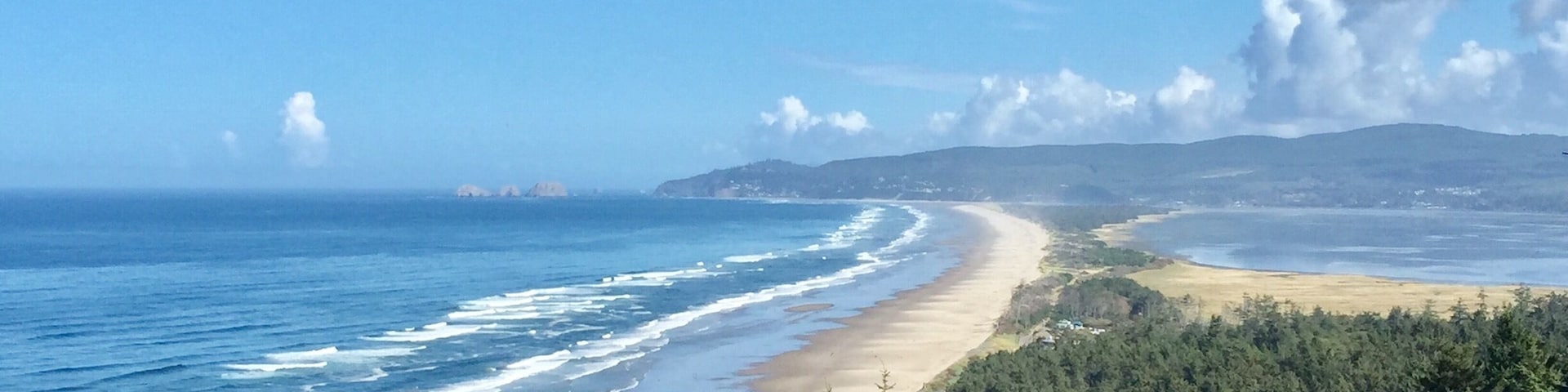 Paisagem costeira com praia de areia, ondas do oceano, floresta densa e colinas distantes.