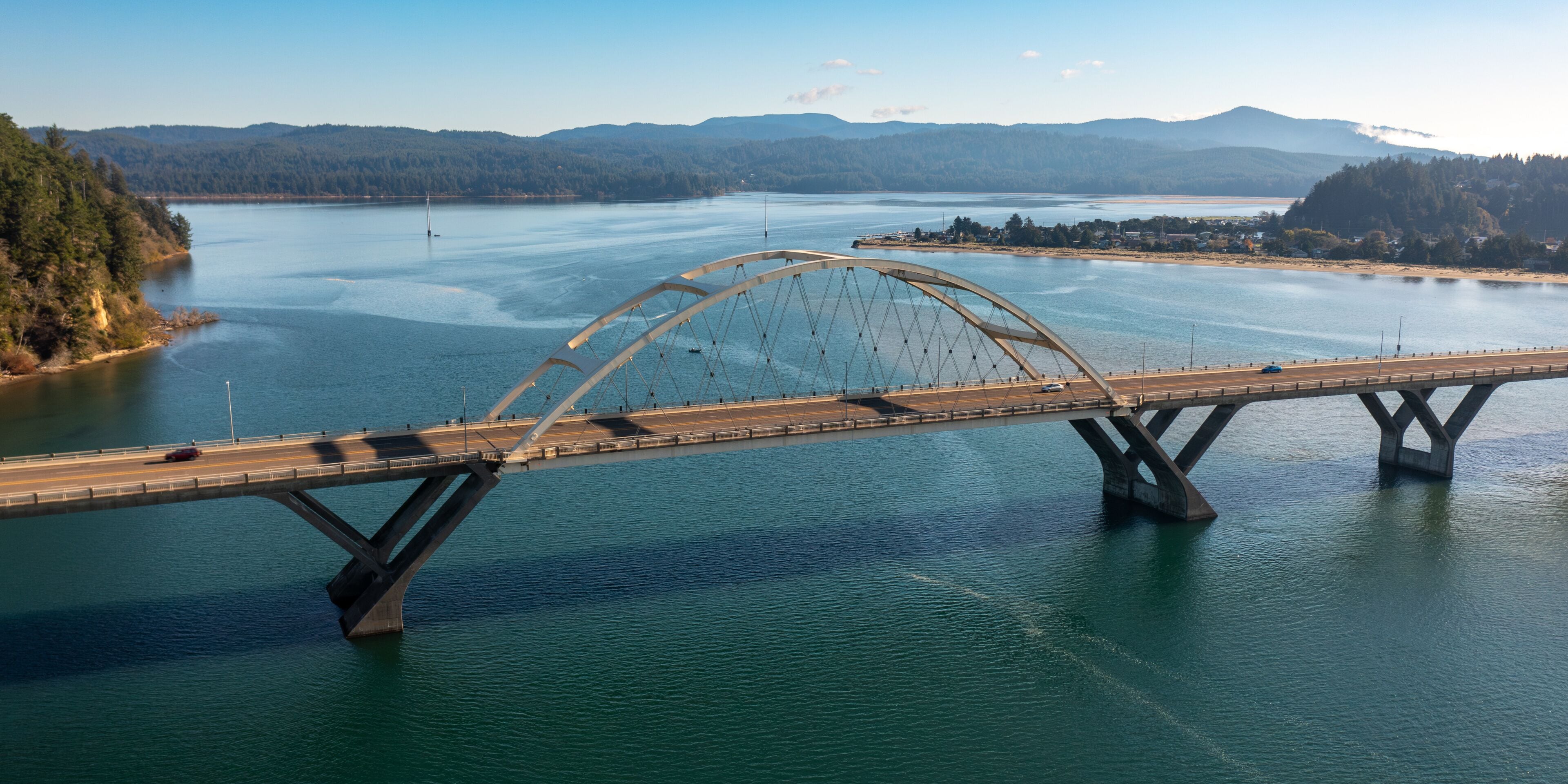 A large suspension bridge spanning a wide river with a forested landscape and mountains in the background.
