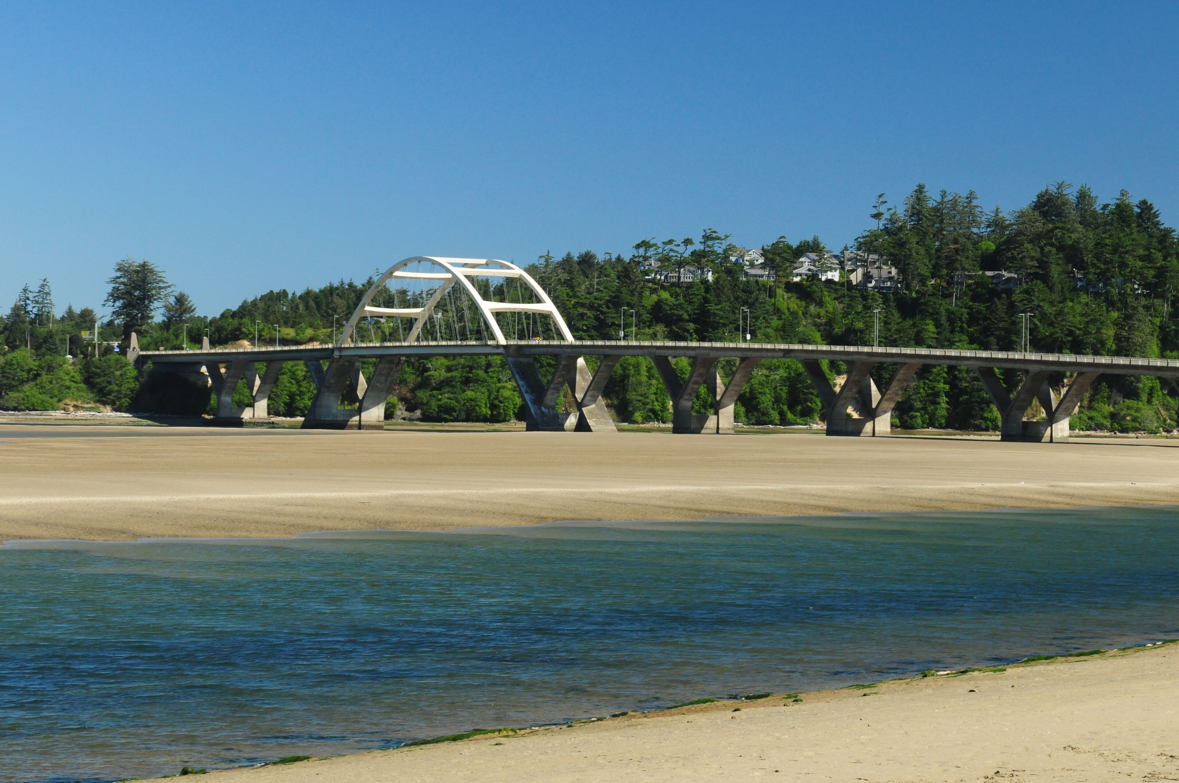 A bridge with a unique arch design spanning a river, surrounded by trees and a clear sky.