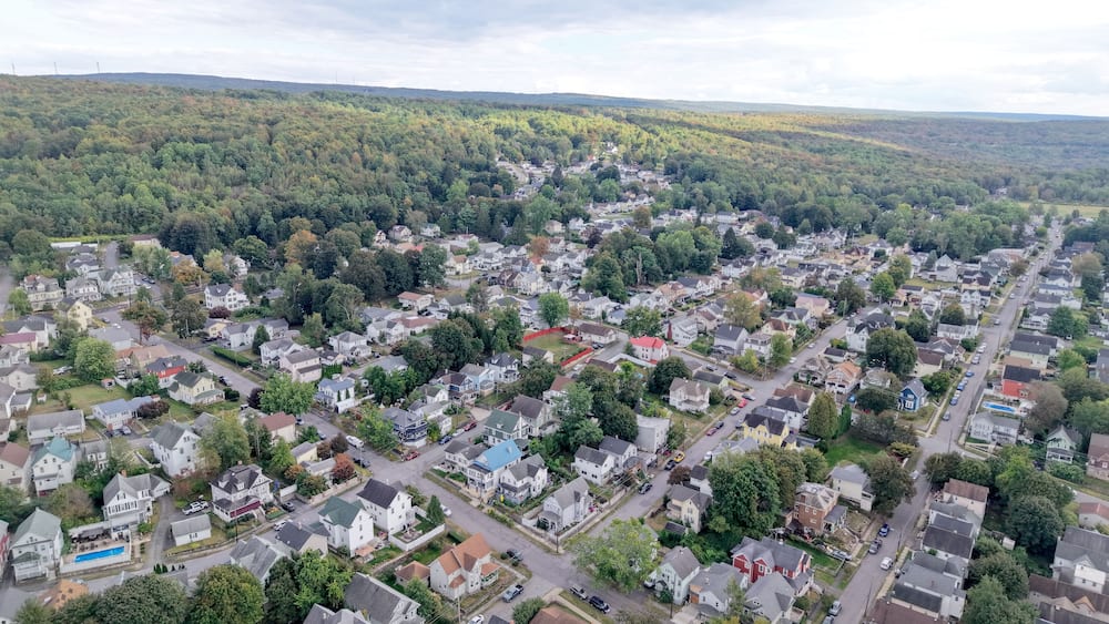 An aerial view of a residential area with houses, streets, and a forested hill in the background.