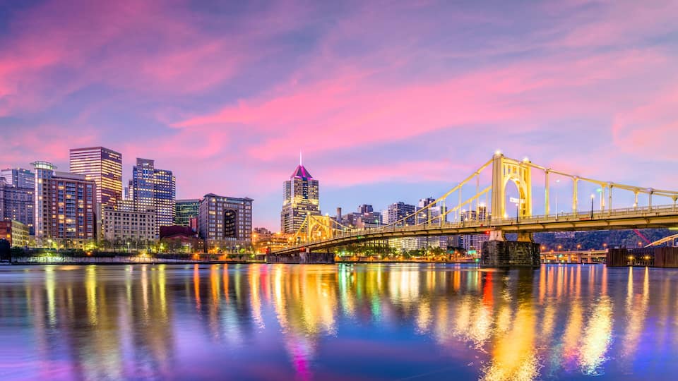 A city skyline with a prominent bridge reflecting in the water, illuminated buildings, and a distinctive skyscraper with a pointed top.