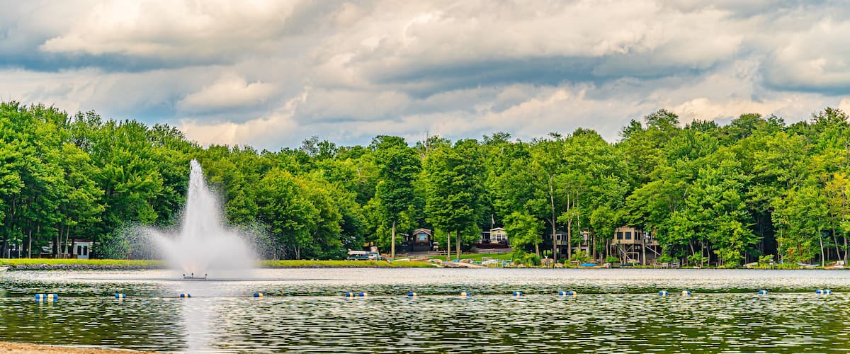 A lake with a fountain, surrounded by dense green trees and houses.