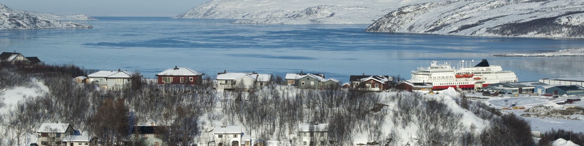 Un pueblo costero nevado con casas, un crucero y colinas cubiertas de nieve.