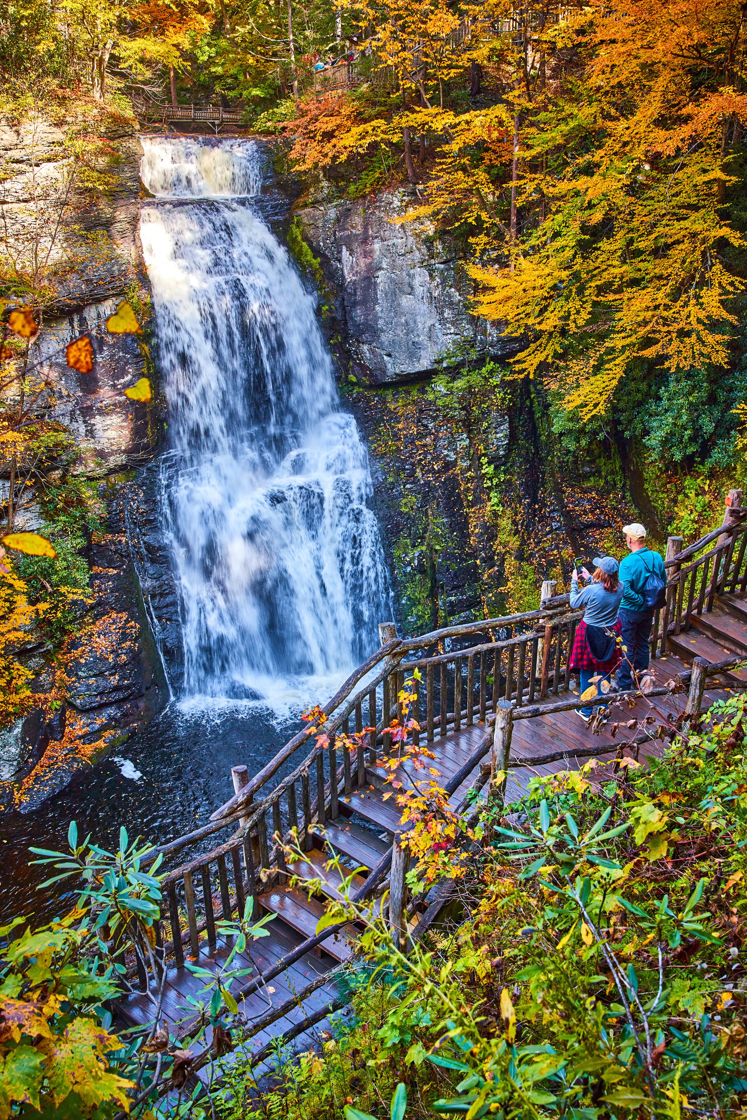 Couple enjoying view from boardwalk of huge stunning waterfall over cliffs surrounded by fall foliage