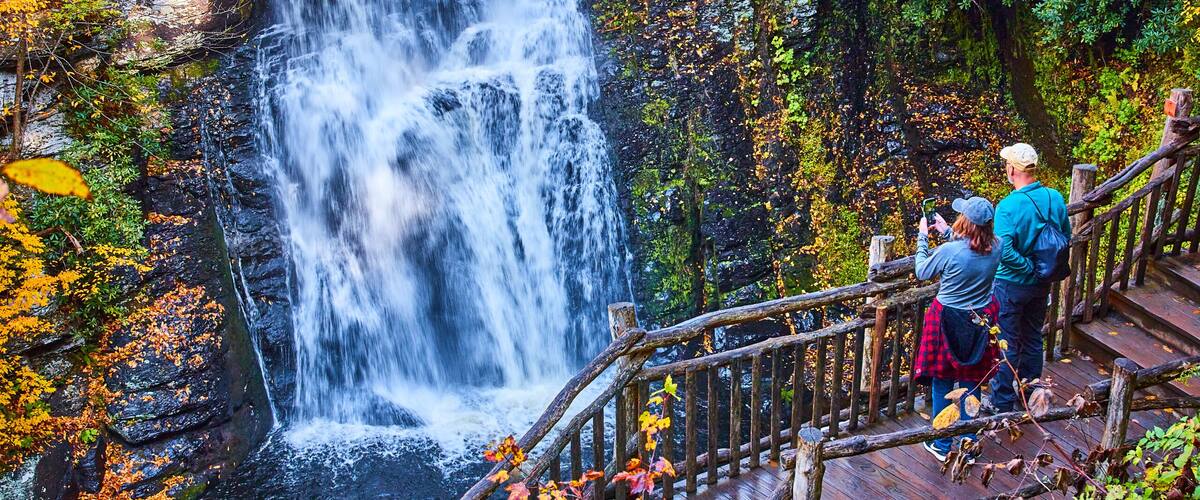Couple enjoying view from boardwalk of huge stunning waterfall over cliffs surrounded by fall foliage