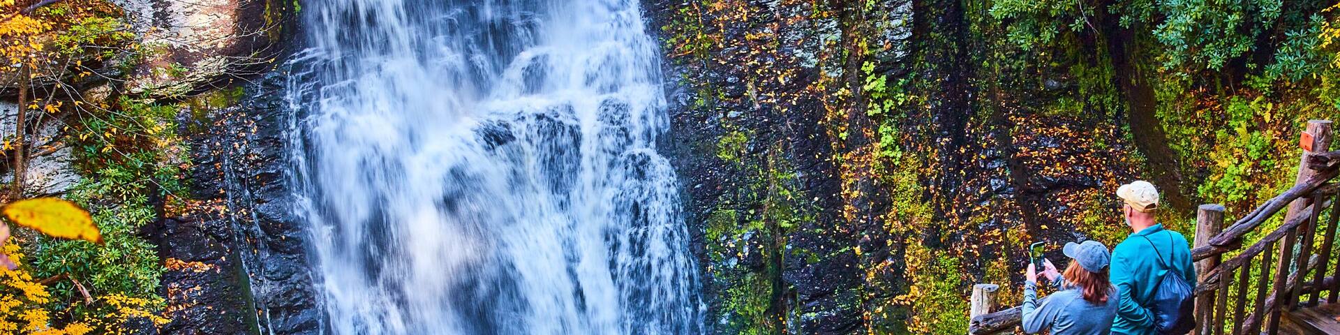 Couple enjoying view from boardwalk of huge stunning waterfall over cliffs surrounded by fall foliage