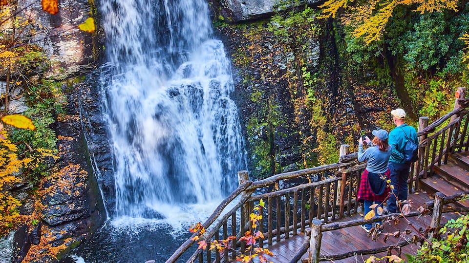 Couple enjoying view from boardwalk of huge stunning waterfall over cliffs surrounded by fall foliage