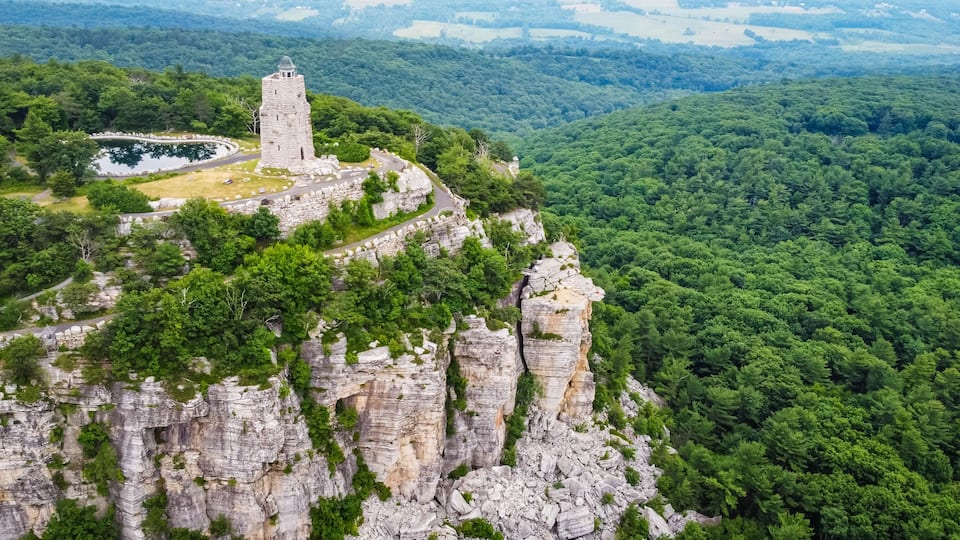 A stone tower on a cliff, surrounded by dense forest and a winding road.