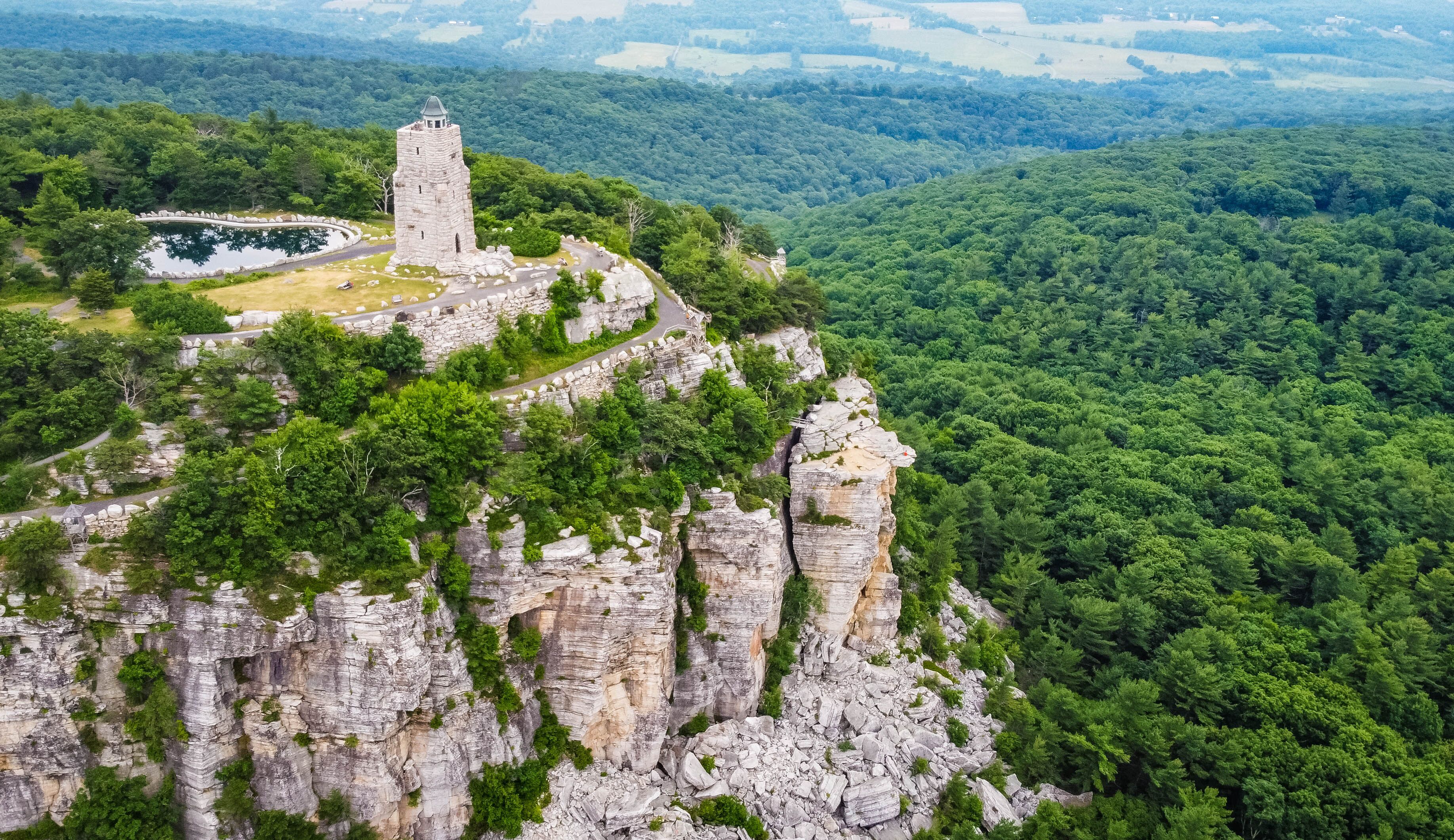 A stone tower on a cliff, surrounded by dense forest and a winding road.